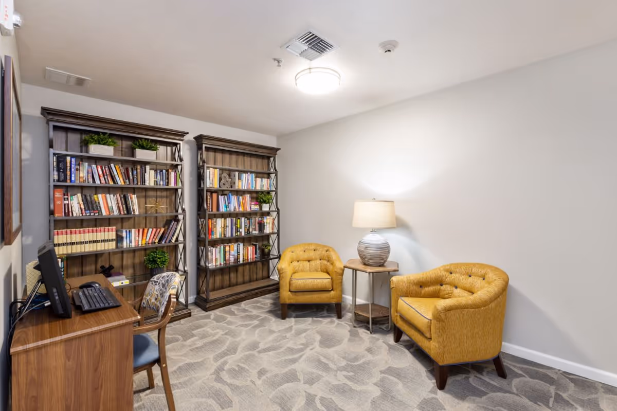 A cozy reading and computer area in a senior living facility featuring two wooden bookshelves filled with books and decorative plants, a wooden desk with a computer and keyboard, a chair with a patterned cushion, two mustard yellow upholstered armchairs, a small round side table with a lamp, and a carpeted floor with a subtle pattern.