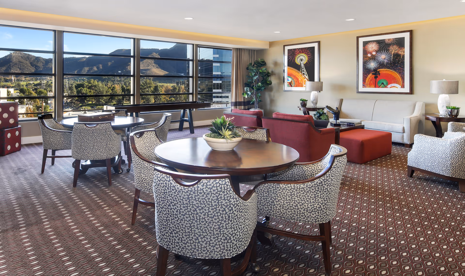 A spacious and well-lit living room area with large windows offering a scenic view of mountains and greenery. The room features round wooden tables with patterned chairs, a shuffleboard table near the windows, a beige sofa with red ottomans, and two framed pictures of fireworks on the wall. There are also decorative plants and table lamps adding to the cozy ambiance.