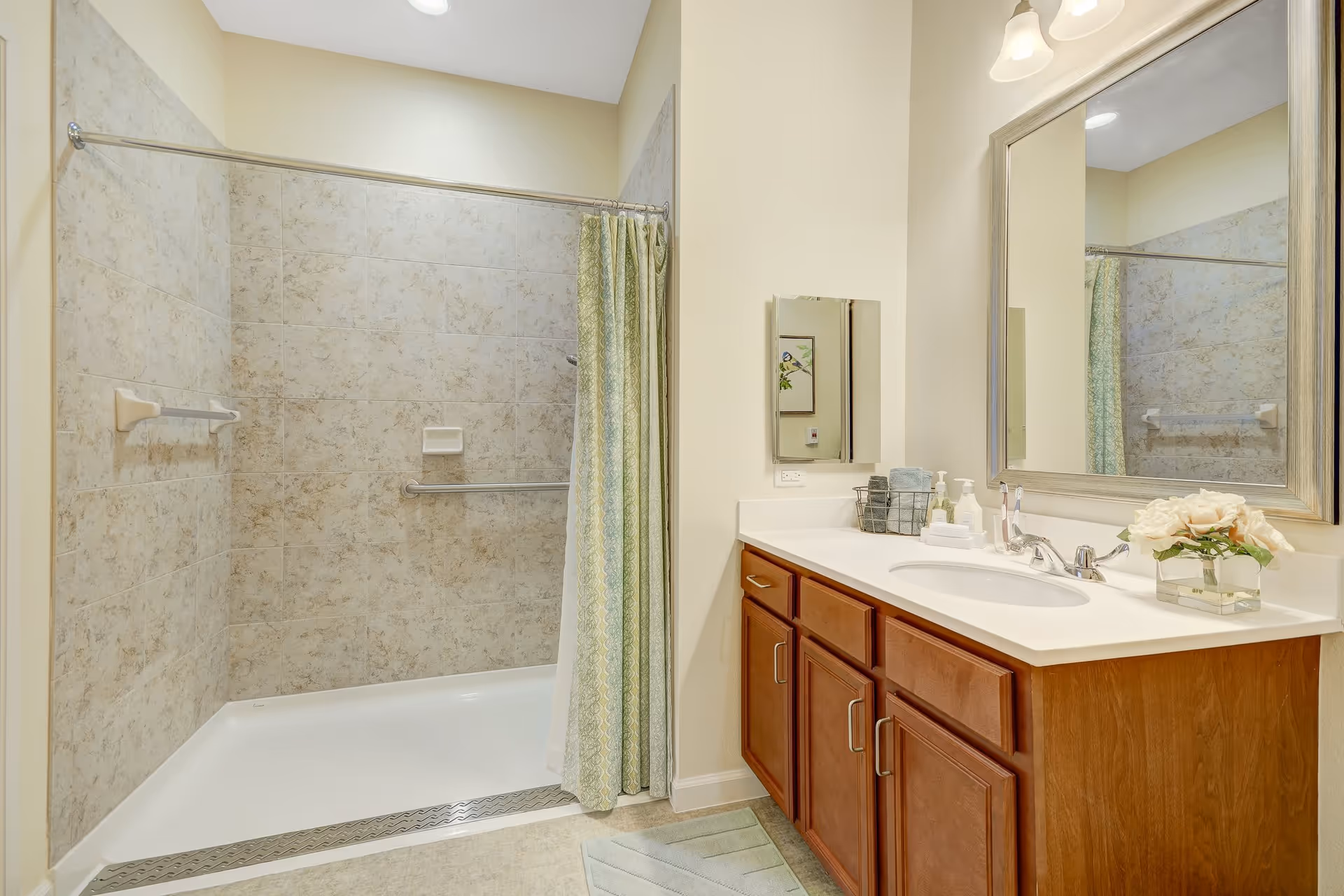 A clean and well-lit bathroom featuring a walk-in shower with beige tiled walls and a green patterned shower curtain. There is a wooden vanity with a white countertop, a sink, a large framed mirror, and various toiletries including soap dispensers, toothbrushes, and a small vase with white flowers.