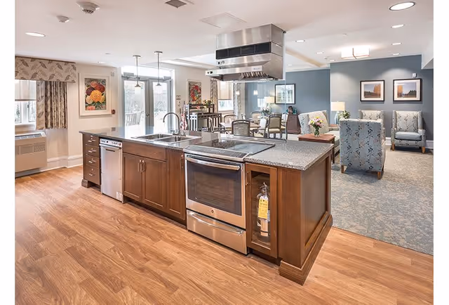 A spacious senior living facility interior featuring a modern kitchen island with a stove, oven, sink, and dishwasher. The kitchen island has wooden cabinetry and a granite countertop. In the background, there is a dining area with tables and chairs, and a comfortable seating area with armchairs and framed artwork on the walls. The flooring is a combination of wood and carpet, and the room is well-lit with recessed ceiling lights.