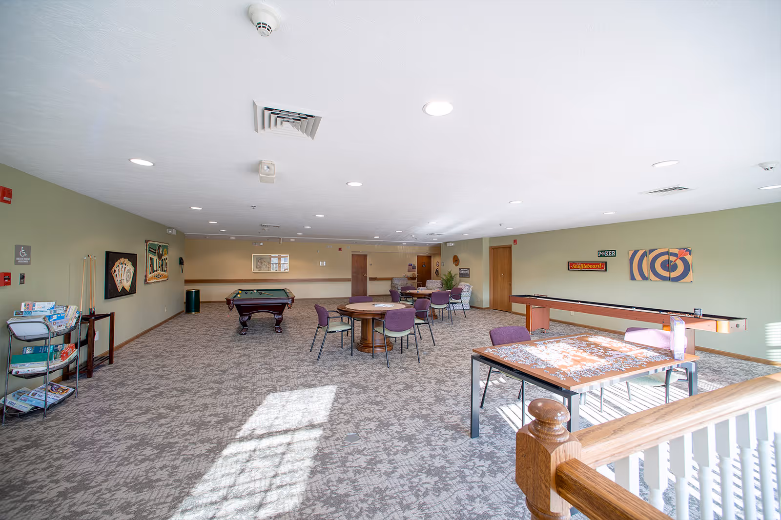 A spacious recreational room with carpeted floor and light green walls. The room contains several tables with chairs, a pool table, a shuffleboard, and a puzzle in progress on one table. There are framed pictures on the walls and a rack with board games and puzzles. The room is well-lit with ceiling lights and natural light coming through windows.