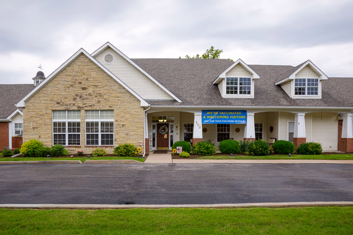 Front exterior view of a single-story assisted living facility building with a stone and siding facade, large windows, a covered entrance with white columns, and a blue banner that reads 'WE'RE VACCINATED & WELCOMING VISITORS ASK OUR TEAM FOR MORE DETAILS'. There are shrubs and small plants along the front of the building and a paved driveway in front.