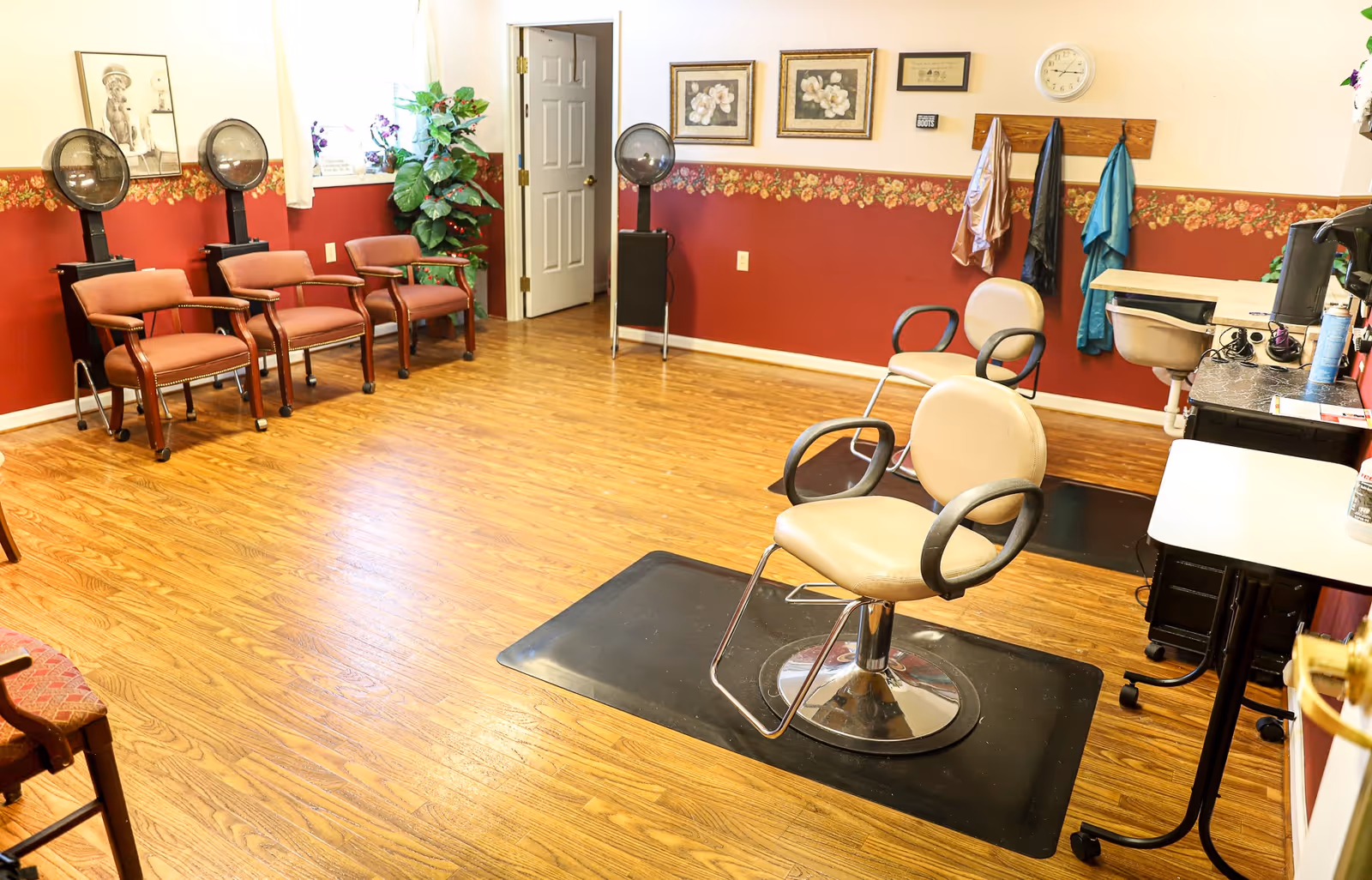 A small salon room with styling chairs, hooded dryers, waiting chairs, and coats hanging on the wall in a senior living facility.
