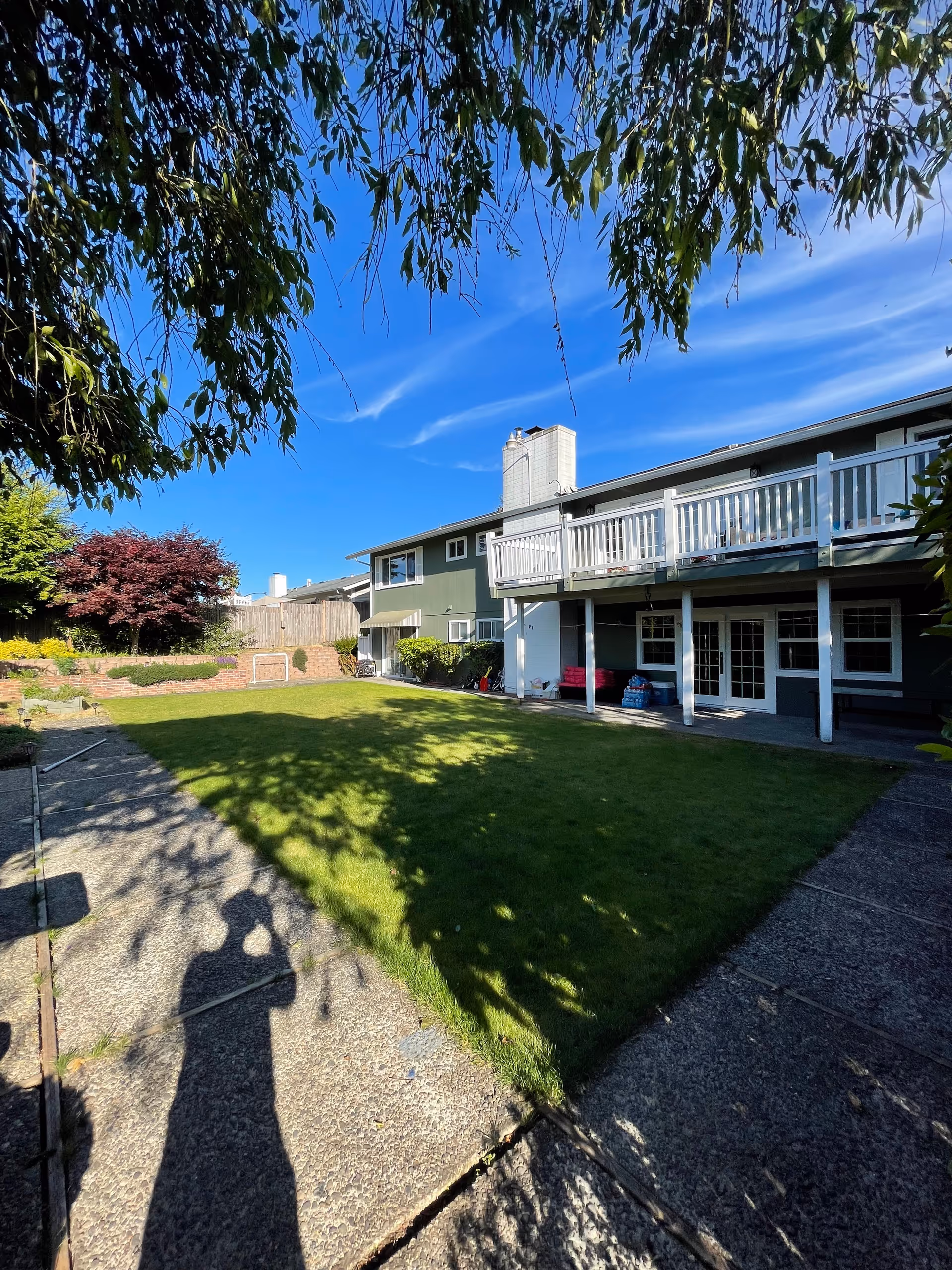 A sunny backyard with a green lawn, concrete paths, and a two-story house featuring an upper deck and patio under a blue sky.