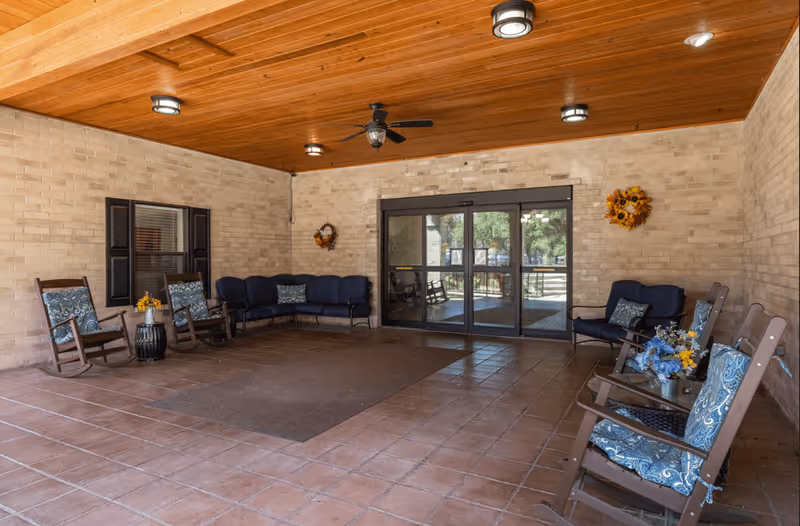 Covered outdoor seating area with a wooden ceiling, ceiling fan, and brick walls. The space includes several rocking chairs with blue patterned cushions, a blue cushioned bench, small tables with flower arrangements, and glass double doors leading outside. Two decorative wreaths hang on the walls.