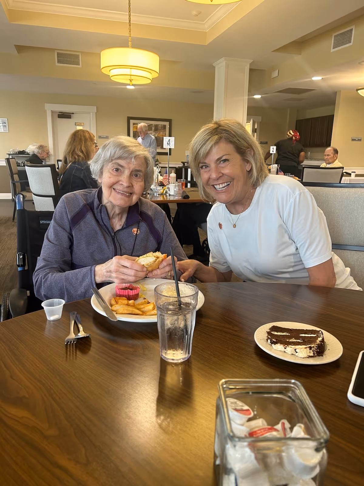 Two women smiling and sitting at a dining table in a senior living facility. One woman is elderly and holding a sandwich with a plate of fries and a cupcake in front of her. The other woman is younger and wearing a white shirt. In the background, other residents and staff are visible in the dining area.