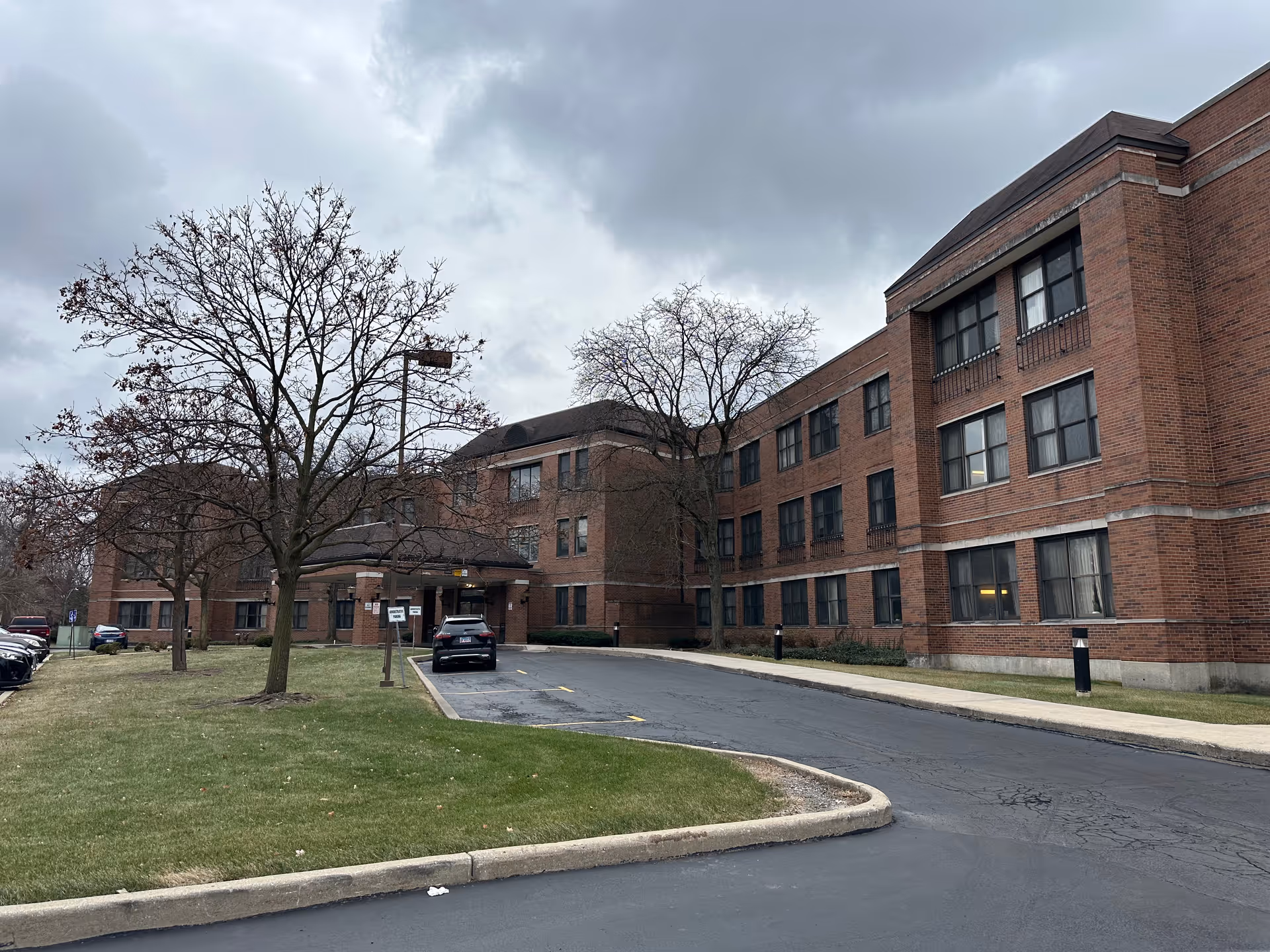 Exterior view of a large brick building with multiple windows, a driveway leading to a covered entrance, several leafless trees, parked cars, and a cloudy sky overhead.