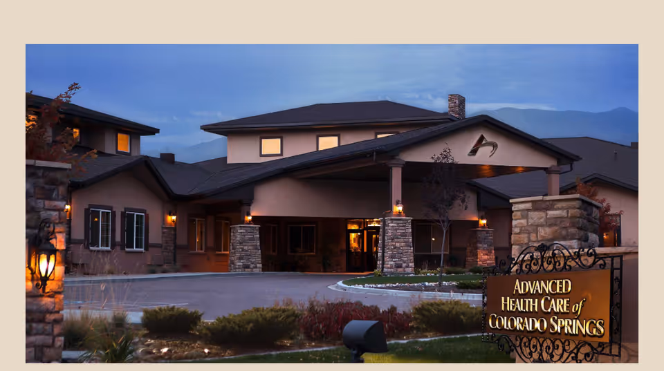 Front entrance of the Advanced Health Care of Colorado Springs building at dusk with illuminated lamps, stone pillars, and a sign in the foreground.