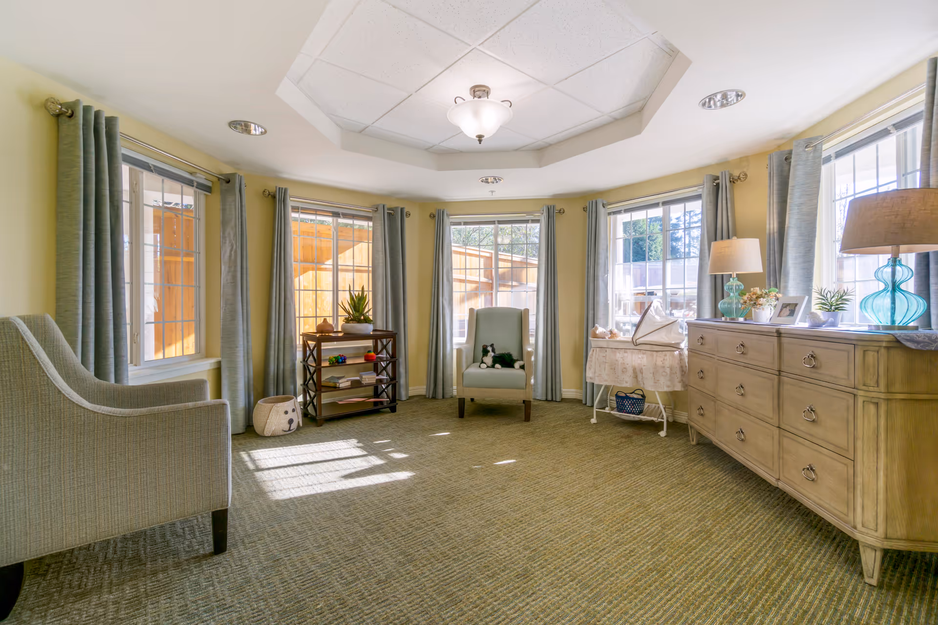 A bright and cozy living room with large windows covered by light blue curtains, two armchairs, a wooden dresser with lamps and decorative items, a small shelf with plants and books, and a carpeted floor. Sunlight streams through the windows, illuminating the room.