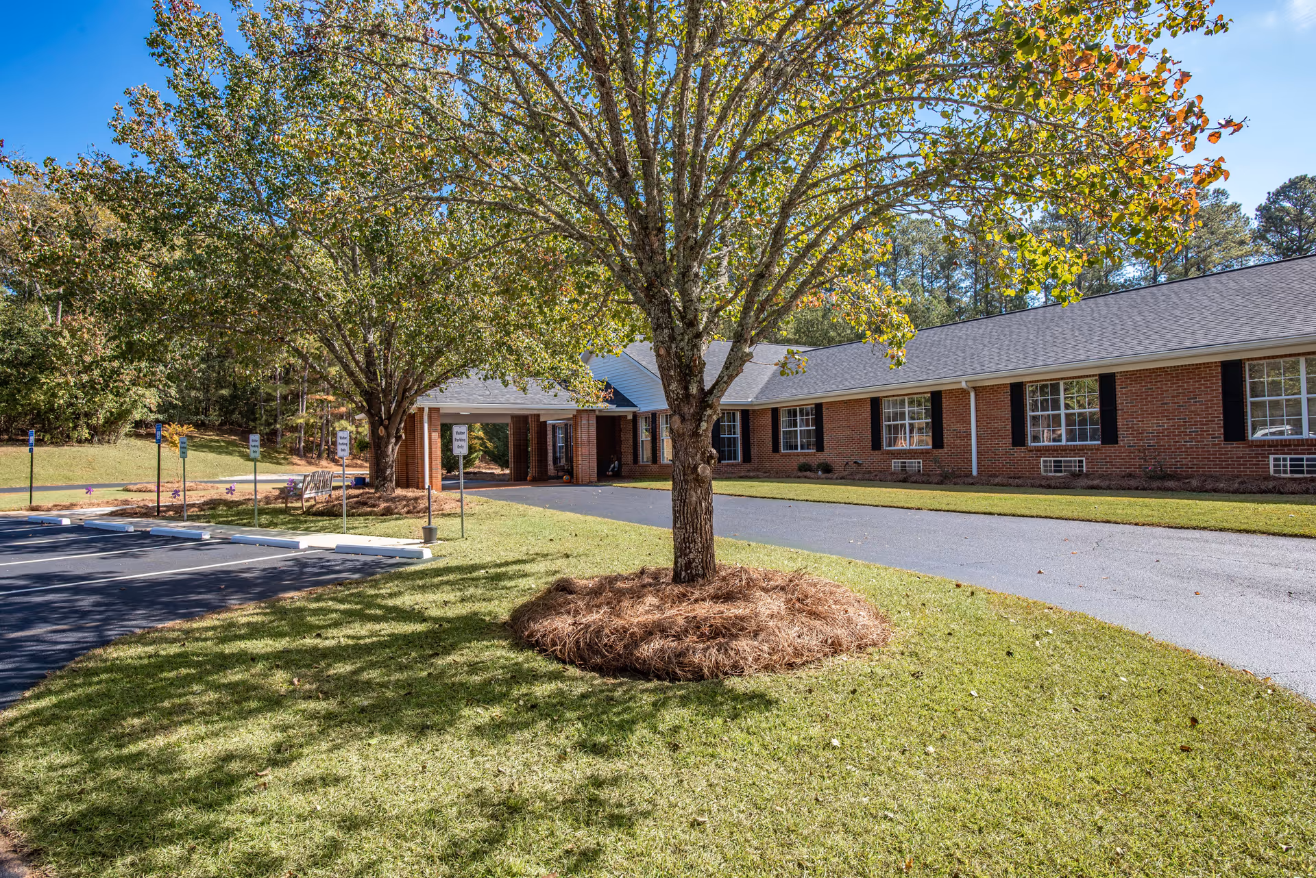 Exterior view of a single-story brick building with a covered entrance, surrounded by green grass and trees under a clear blue sky. There is a paved driveway and parking spaces with signs indicating visitor parking only.