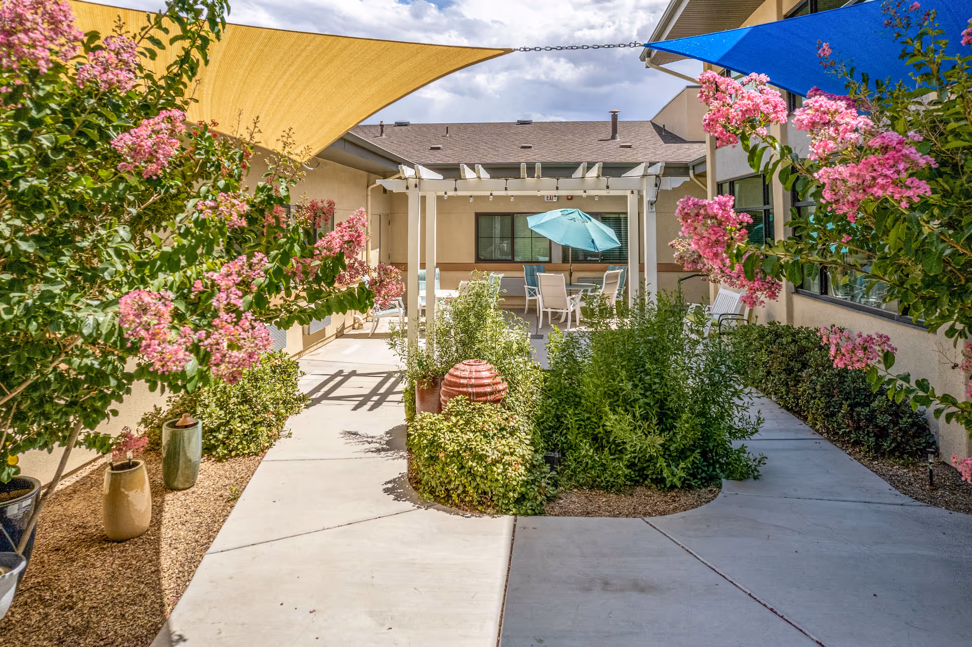 Outdoor courtyard area at The Meadows at Escalante featuring a concrete walkway surrounded by green plants and pink flowering bushes. There are colorful shade sails overhead in yellow and blue, and a white pergola with patio furniture and a turquoise umbrella in the background.