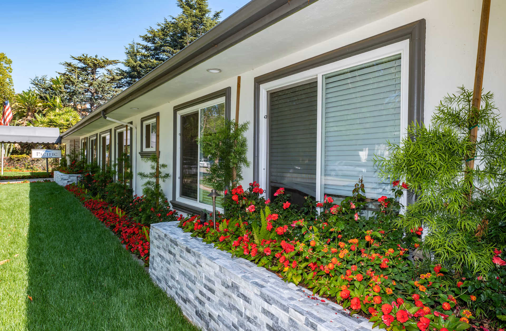 Exterior view of a single-story building with large windows and a well-maintained garden bed filled with red flowers and green plants. The building has a white facade with gray trim around the windows. There is a green lawn in front and trees in the background. A sign near the entrance reads 'Pacific Post-Acute'.