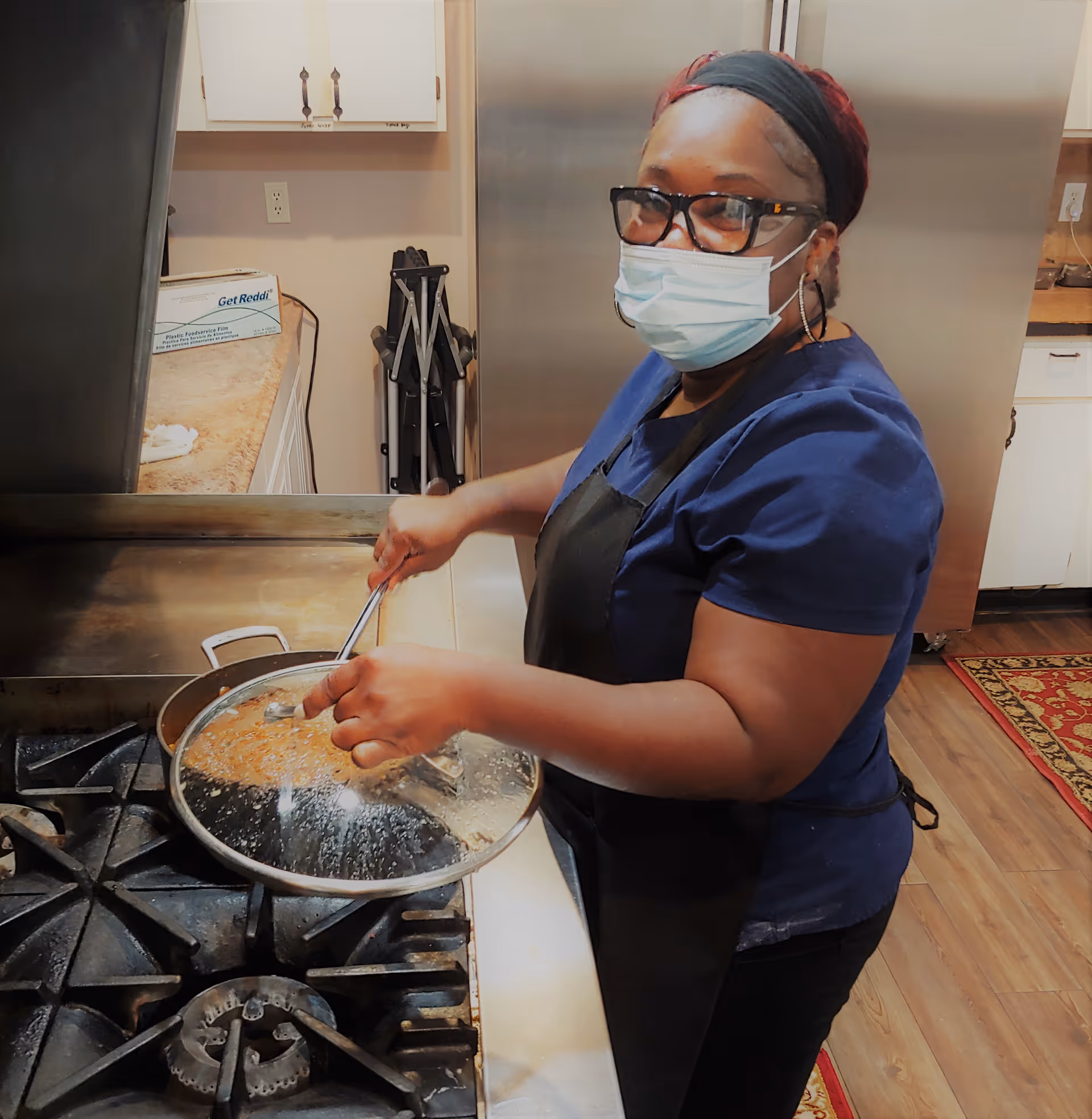 A masked staff member wearing glasses and an apron stirs food in a pan on a commercial stove in a kitchen.