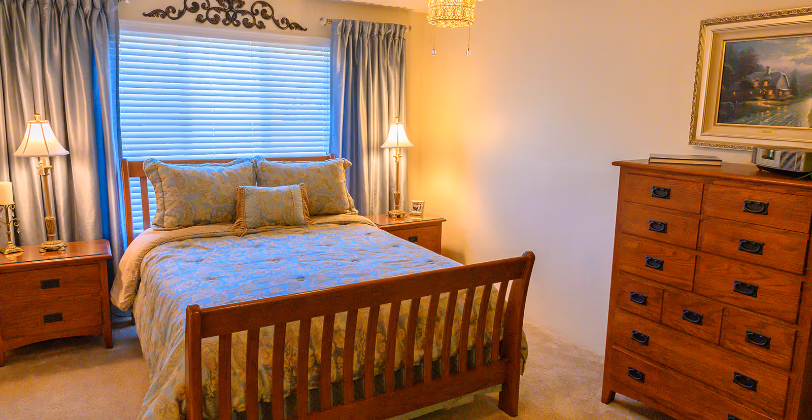 A cozy bedroom with a wooden bed frame and matching wooden nightstands on either side. The bed is made with a patterned comforter and several pillows. There are two table lamps on the nightstands, and a window with closed blinds and curtains behind the bed. A wooden dresser with multiple drawers and a framed painting above it is on the right side of the room.