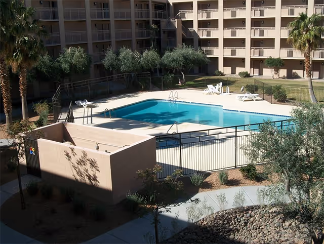 Outdoor swimming pool area surrounded by a black metal fence with lounge chairs and palm trees, located within a multi-story residential building complex.