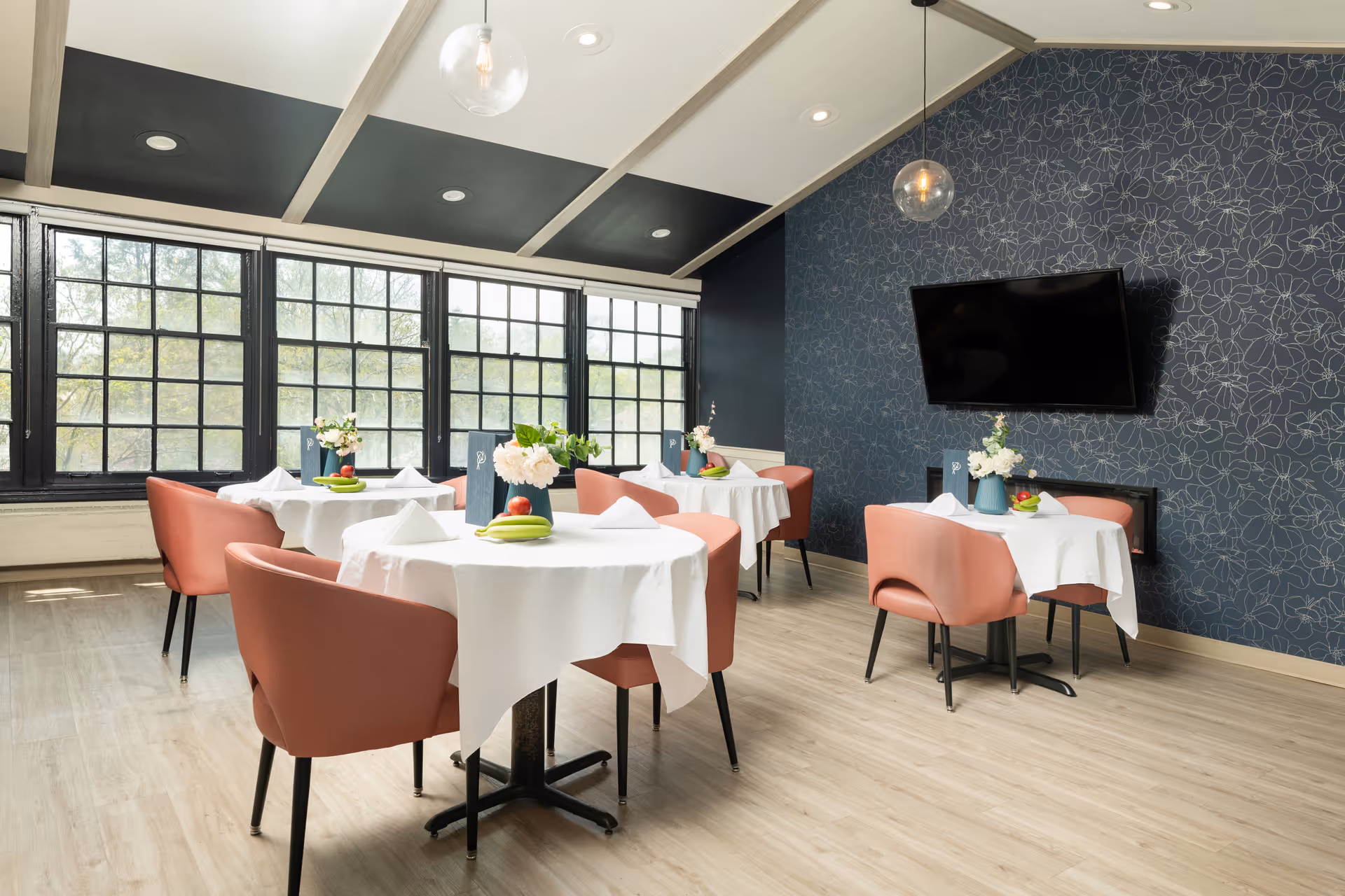 A bright dining room with four round tables covered in white tablecloths, each set with napkins, plates, and small floral centerpieces. The room features large black-framed windows letting in natural light, pink upholstered chairs, light wood flooring, a dark blue floral patterned accent wall, a mounted flat-screen TV, and modern pendant lights hanging from a white ceiling with exposed beams.