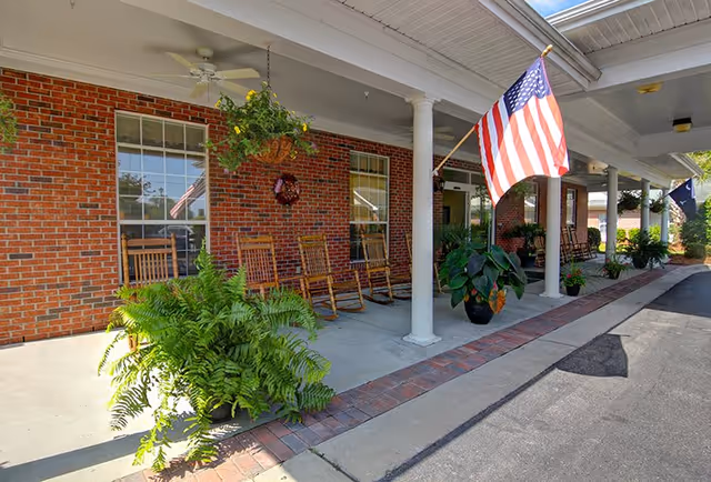 Covered porch area of a brick building with several wooden rocking chairs lined up against the wall, hanging plants, potted plants, white columns, an American flag, and a ceiling fan.