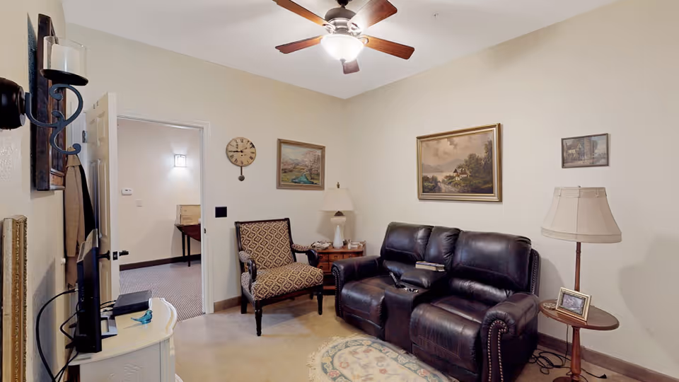 A cozy living room with a dark leather loveseat, a patterned armchair, two side tables with lamps, and framed paintings on beige walls. A ceiling fan with a light is mounted on the ceiling. An open door leads to another room with a table and a wall light.