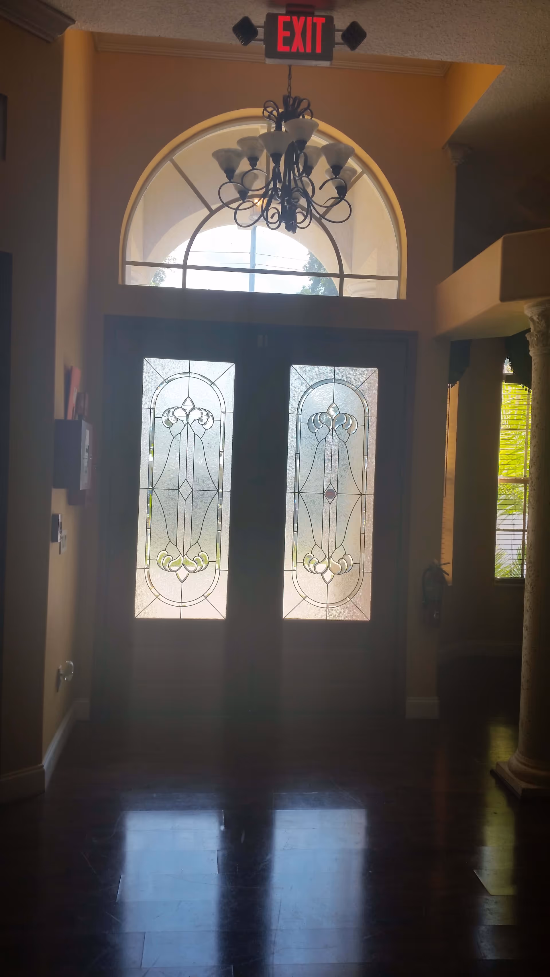 Interior view of a building entrance with double decorative glass doors and an arched window above. A chandelier hangs from the ceiling and an illuminated red EXIT sign is mounted above the doors. The floor is dark wood and there is a column visible on the right side.