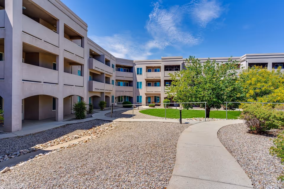 Courtyard with a winding walkway leading to a three-story senior living building with balconies and landscaped trees under a blue sky.