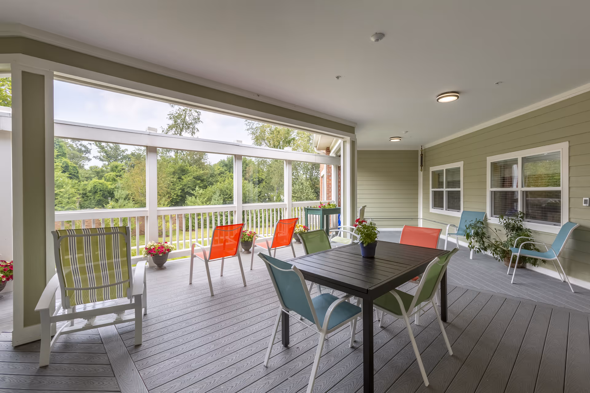 Covered outdoor patio area with a black table surrounded by colorful chairs, additional chairs along the sides, potted plants on the floor and table, and a view of green trees and grass beyond the white railing.