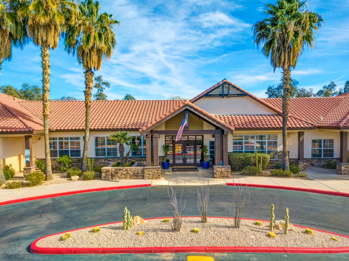 Front exterior view of The Auberge at Scottsdale facility with a tiled roof, stone accents, an entrance with a covered porch, and an American flag. The driveway curves around a landscaped area with desert plants and palm trees.