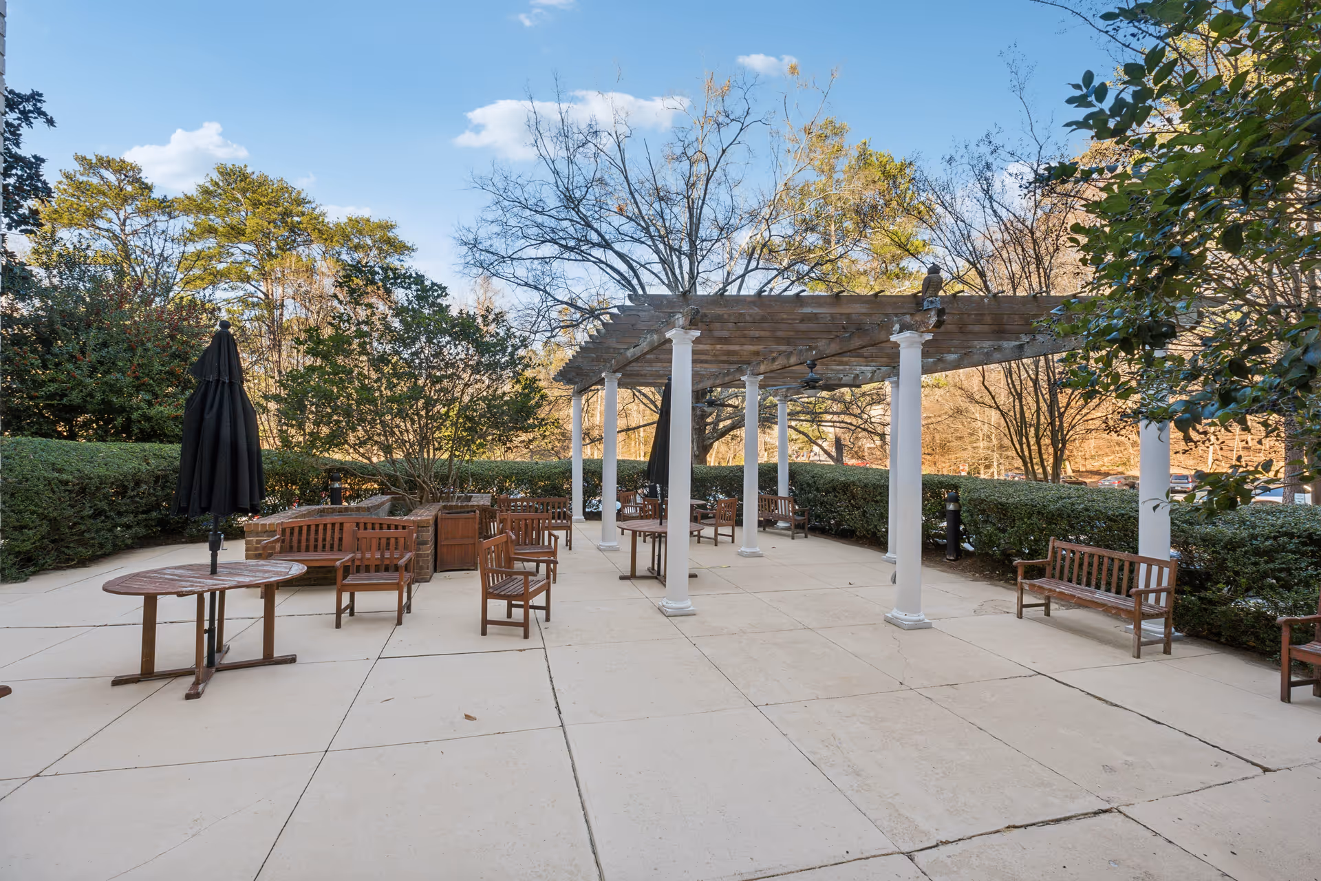 Outdoor patio area with wooden benches, tables, and chairs arranged on a concrete surface. A pergola with white columns provides partial shade. Surrounding the patio are green bushes and trees under a blue sky with some clouds.