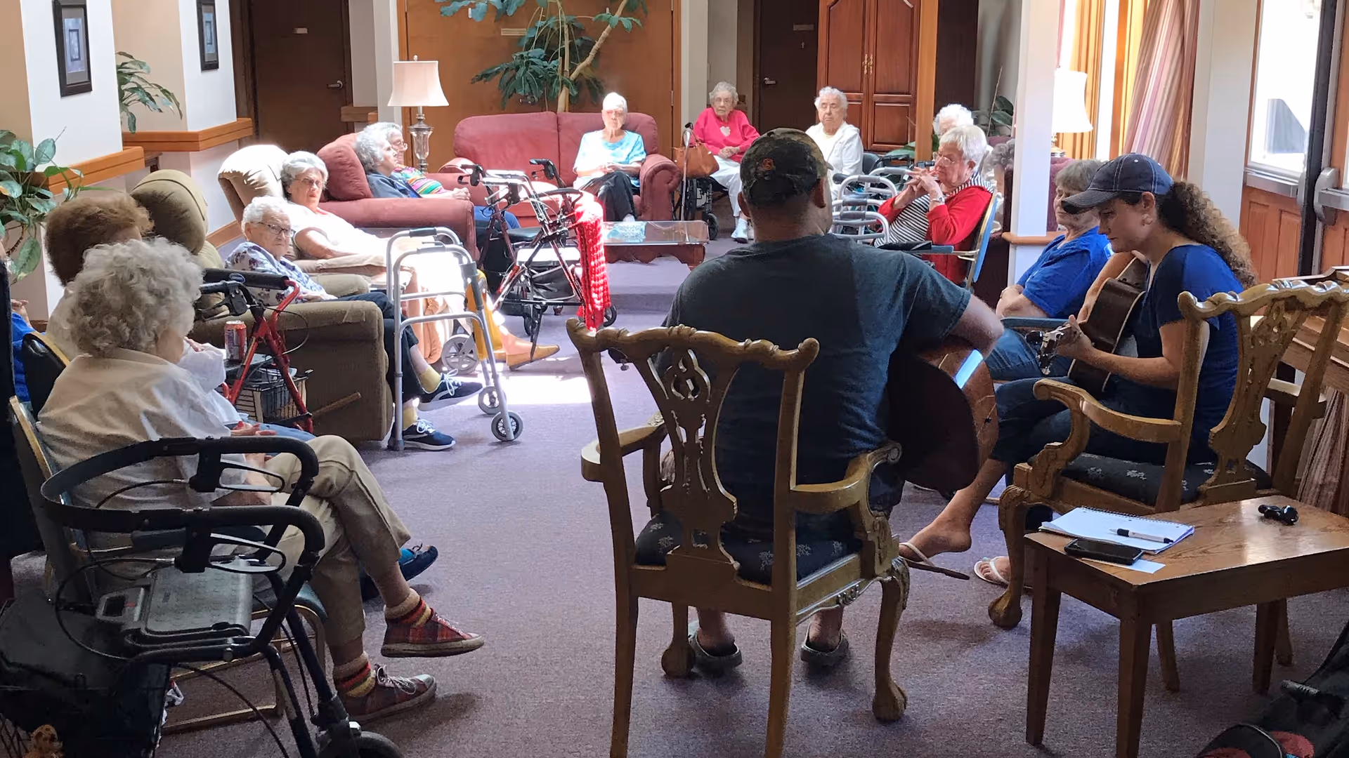 A group of elderly people seated in a living room area, some in wheelchairs and others in armchairs, attentively listening to two individuals playing guitars. The room is well-lit with natural light from windows and decorated with plants, lamps, and framed pictures on the walls.