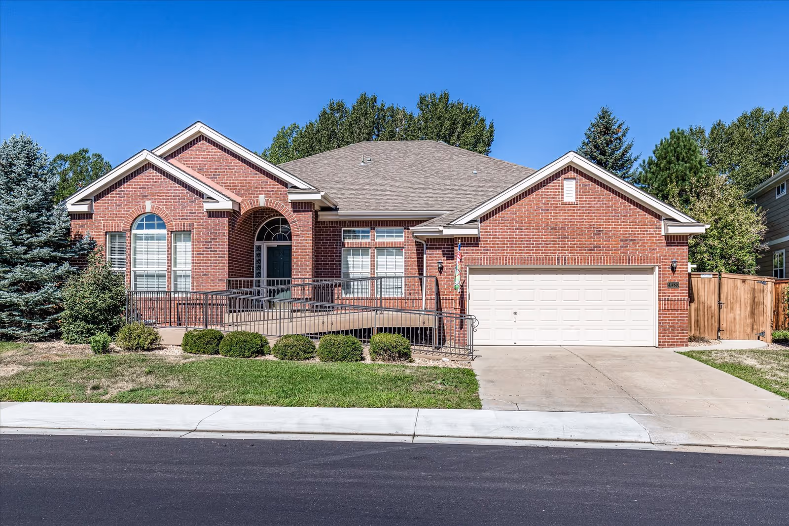 Front exterior view of a single-story brick house with a gray shingled roof, a white garage door, a wheelchair accessible ramp leading to the front door, and neatly trimmed bushes and trees in the yard under a clear blue sky.