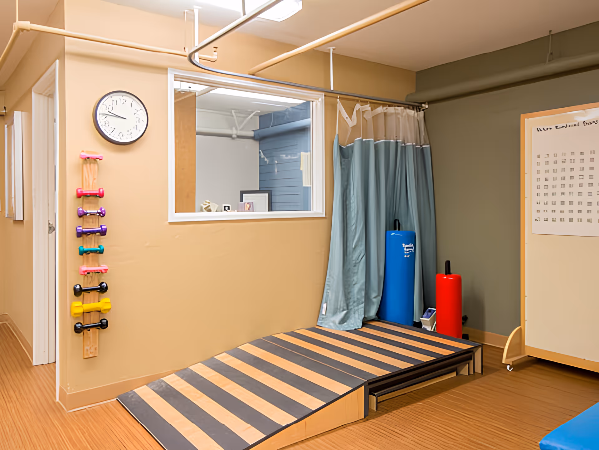 A therapy or exercise room with a wooden ramp, a wall-mounted rack holding colorful small dumbbells, a clock on the wall, a window looking into another room, a blue exercise mat rolled up, a red cylindrical cushion, and a curtain partially drawn to the side.