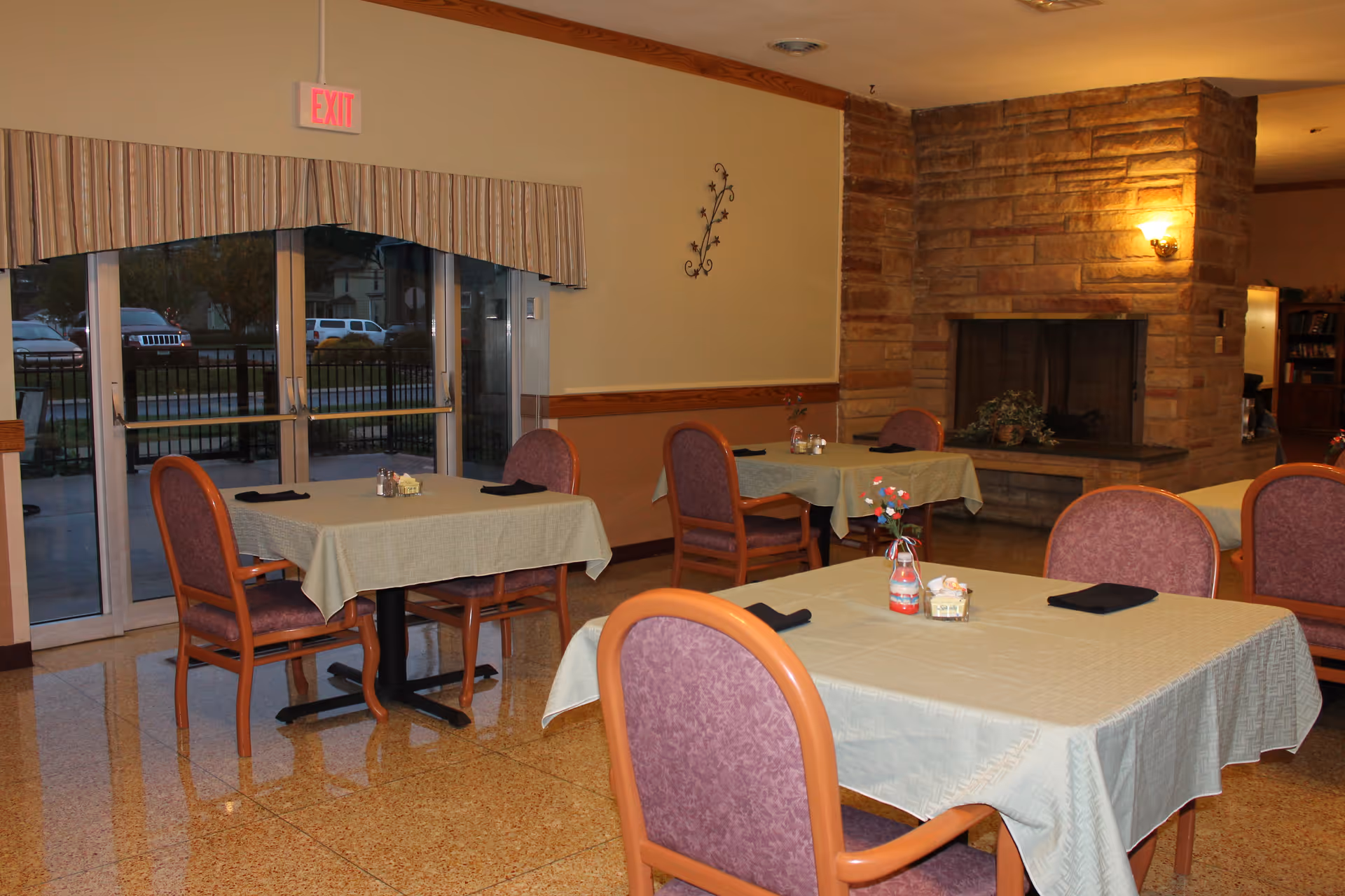 Dining area with tables covered in beige tablecloths and purple cushioned chairs. Each table has a small centerpiece and black napkins. There is a stone fireplace on the right wall and glass doors leading outside on the left side. The room has a warm, cozy atmosphere with soft lighting.