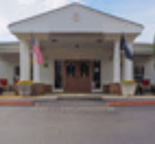 Front entrance of The Gables facility with a covered porch supported by white columns, two flags on either side, and double wooden doors in the center.