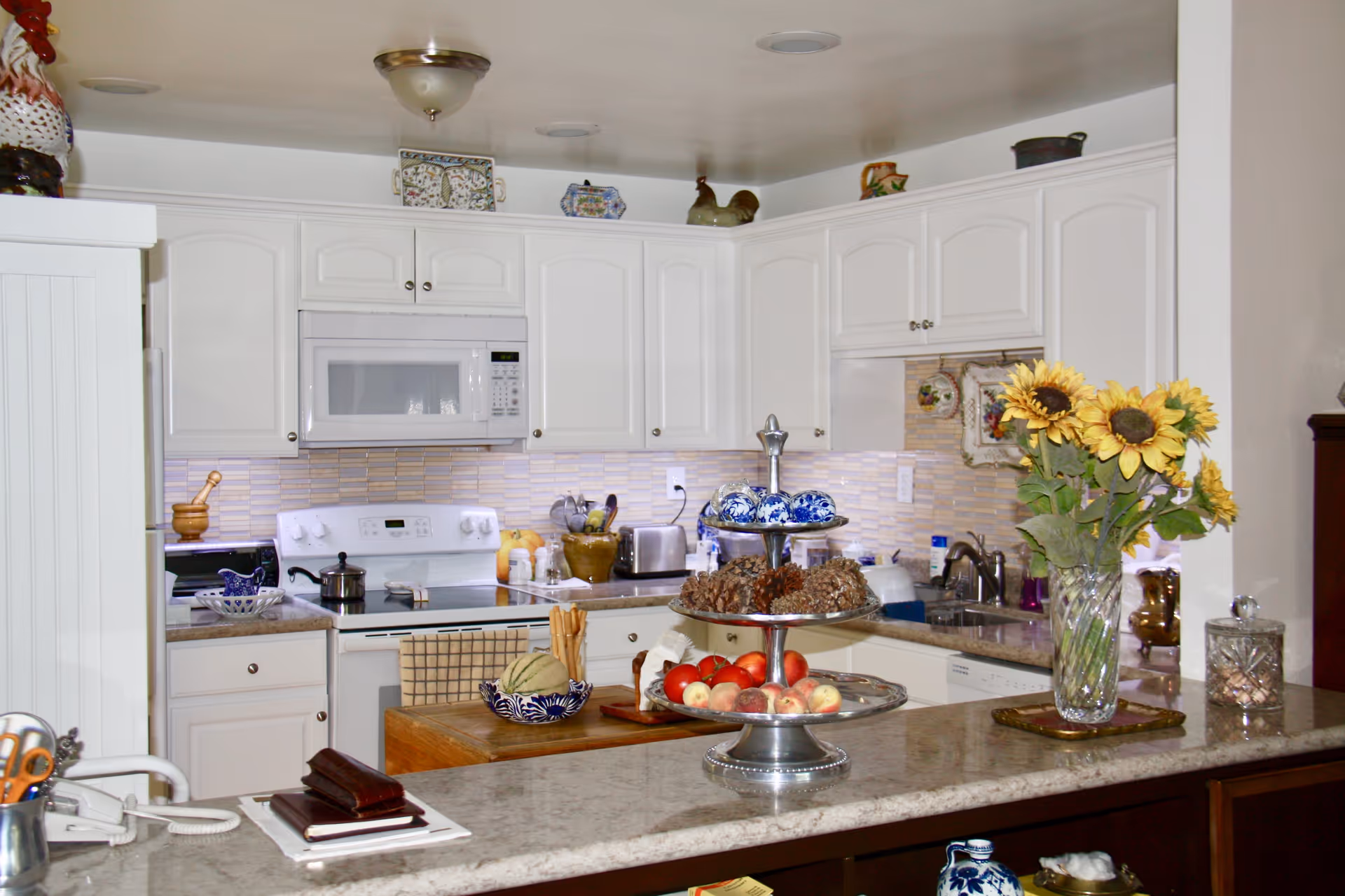 A bright kitchen with white cabinets and a beige tiled backsplash. The kitchen features a white microwave above a white stove, a toaster, and various kitchen utensils on the countertop. A three-tiered tray on the counter holds decorative items and fruit. A vase with sunflowers is placed on the granite countertop in the foreground.
