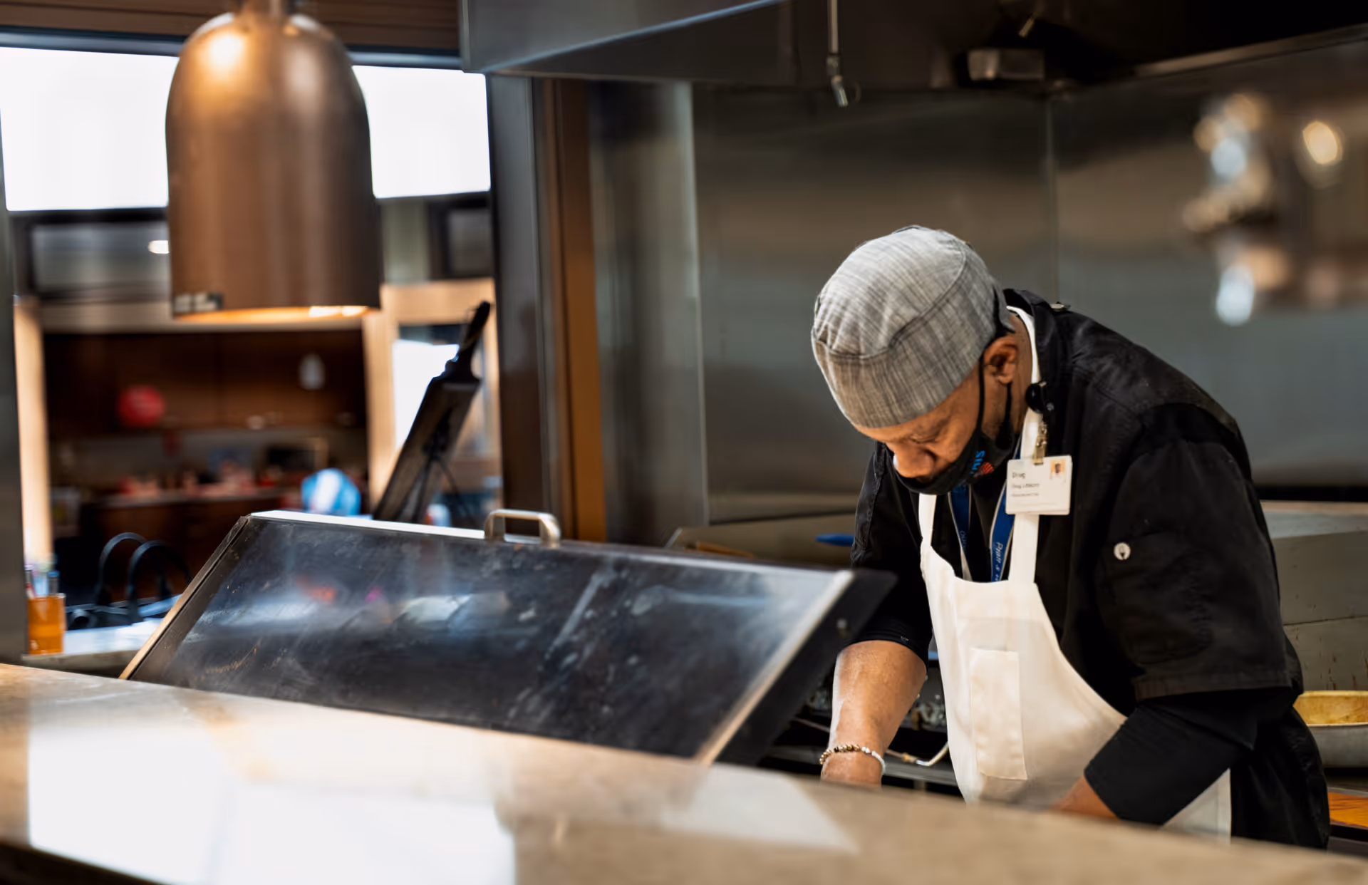 A chef wearing a gray cap, black shirt, and white apron is working in a kitchen, focused on preparing food. The kitchen has stainless steel appliances and a heat lamp above the counter.
