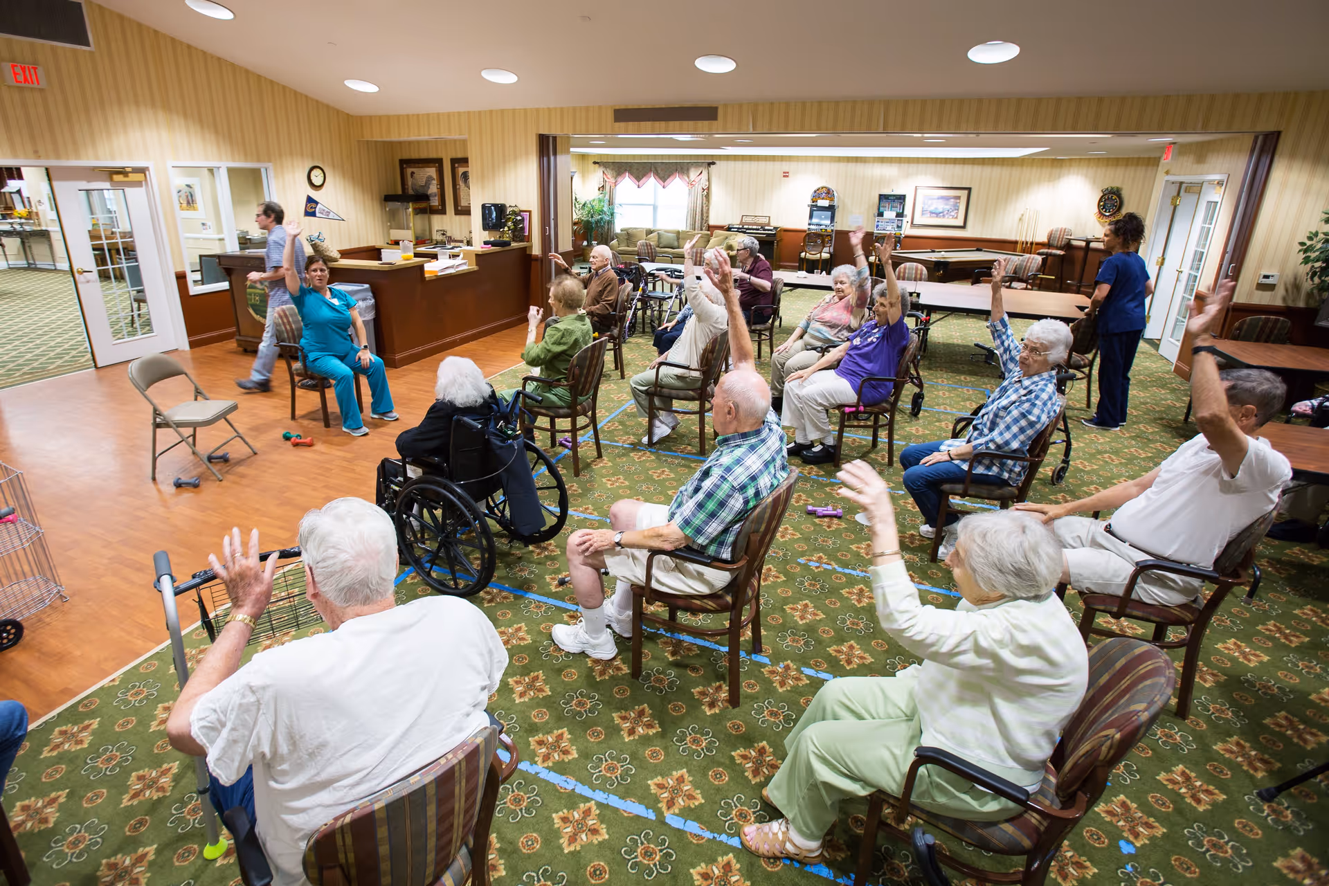 A group of elderly people seated in a circle in a common room participating in a seated exercise session led by a staff member in blue scrubs. The room has a patterned green carpet, wooden flooring near the entrance, and various tables and chairs. Another staff member is standing near the back of the room.
