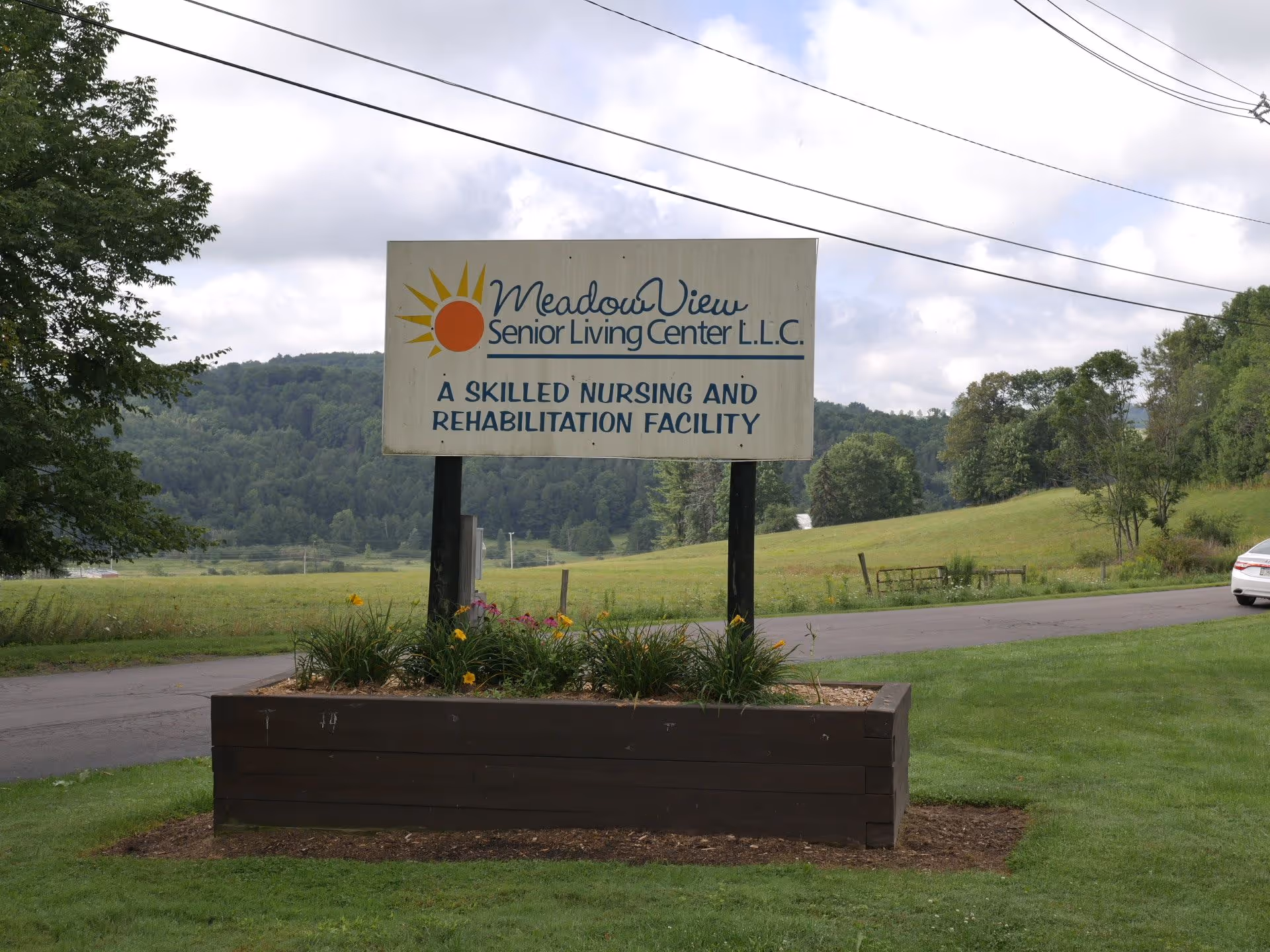 A roadside sign for Meadow View Senior Living Center surrounded by a planter and grassy landscape with hills in the background.