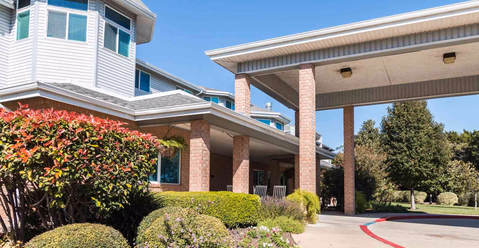 Front entrance of a senior living building with a covered drop-off canopy, brick columns, windows, and landscaped shrubs under a clear sky.