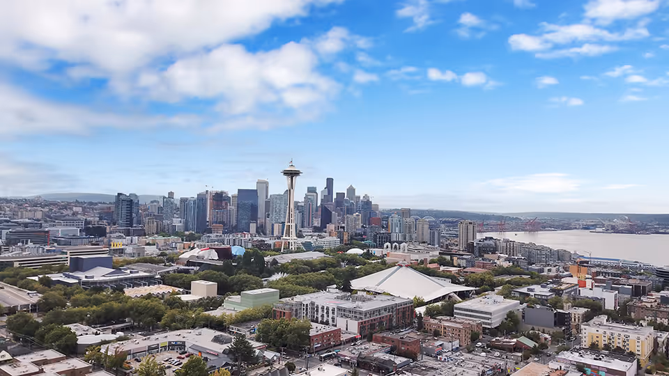 Aerial view of a city skyline featuring the Space Needle prominently in the center, surrounded by numerous buildings, greenery, and a body of water under a partly cloudy blue sky.
