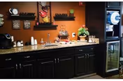 Small kitchenette/coffee station with dark cabinets, a countertop holding a coffee maker, cups, snacks, decorative shelves, and a mini-fridge.