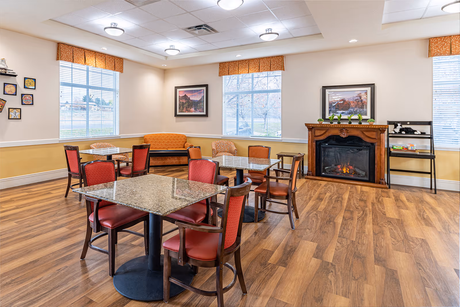 A cozy common area with wooden flooring, two square tables with red cushioned chairs, an orange couch and chair, a wooden fireplace with a fire burning, and large windows with orange valances letting in natural light. The walls are decorated with framed pictures and small plants on the fireplace mantel.