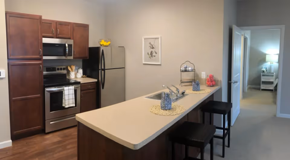 Open kitchen with a long island and bar stools, stainless steel appliances, dark cabinets, and a view into an adjacent bedroom.