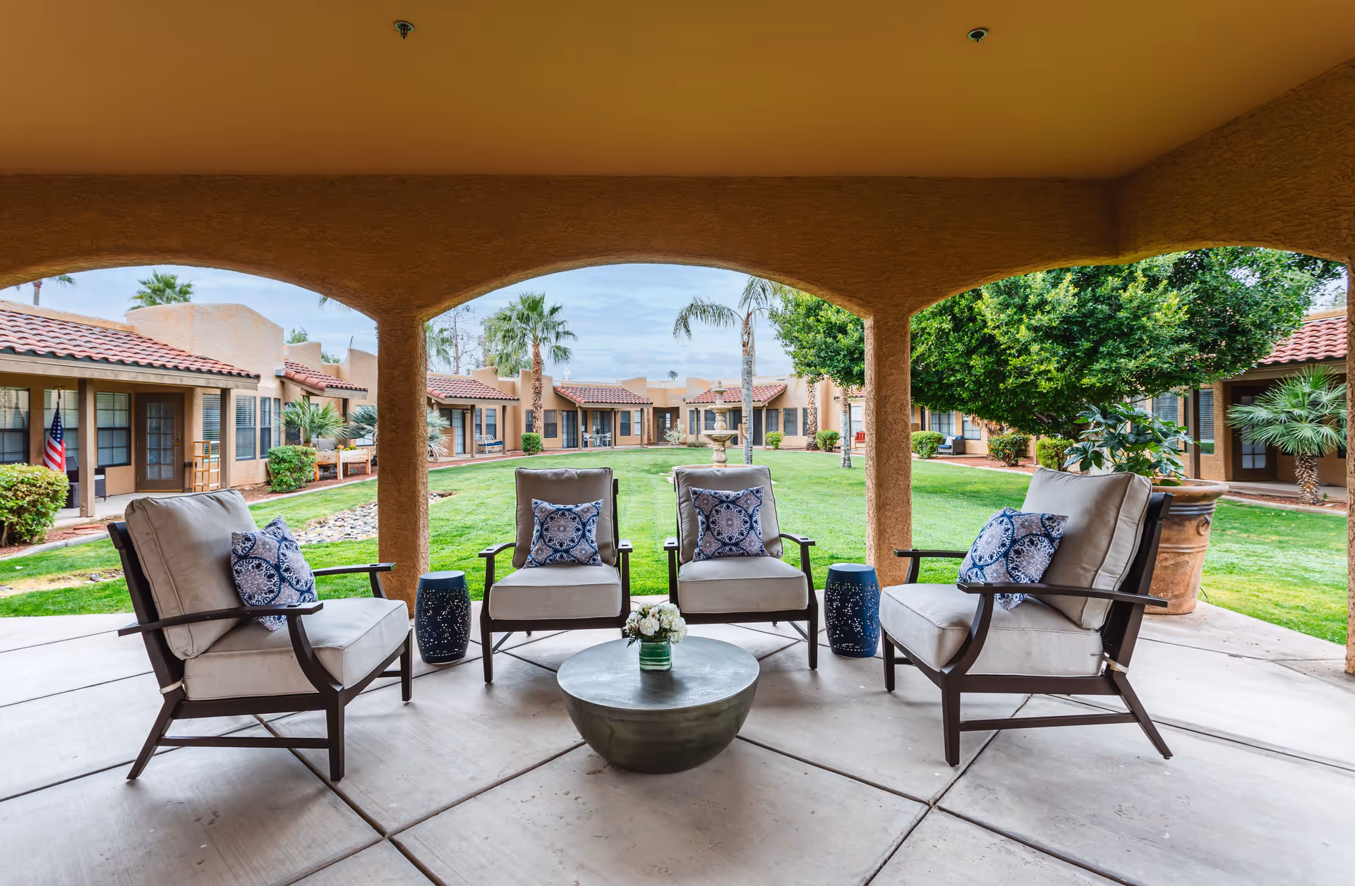 Covered outdoor seating area with four cushioned chairs arranged around a round metal table with a small flower arrangement. The seating area overlooks a green lawn with palm trees, shrubs, and a fountain, surrounded by single-story buildings with red tile roofs.