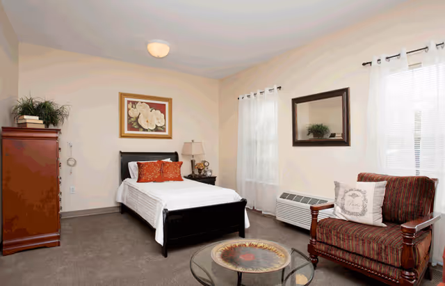 A cozy bedroom with a single bed featuring white bedding and two orange decorative pillows. There is a wooden nightstand with a lamp and clock beside the bed. A wooden dresser with a plant and books on top is against one wall. The room has two windows with white curtains, a striped armchair with a decorative pillow, a glass coffee table with a decorative plate, and a wall mirror above a heating unit.