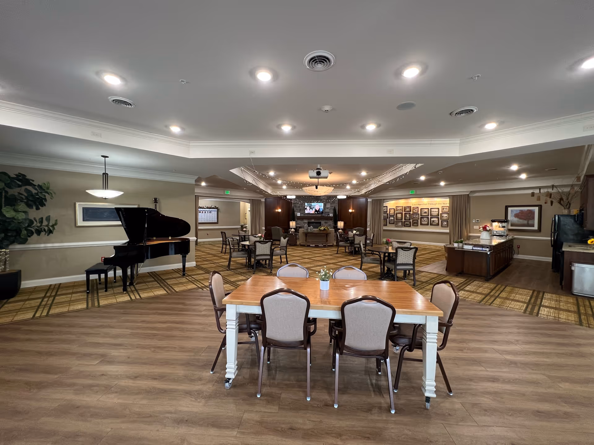Spacious senior living common dining and activity room with tables and chairs, a grand piano, and a TV on the far wall.