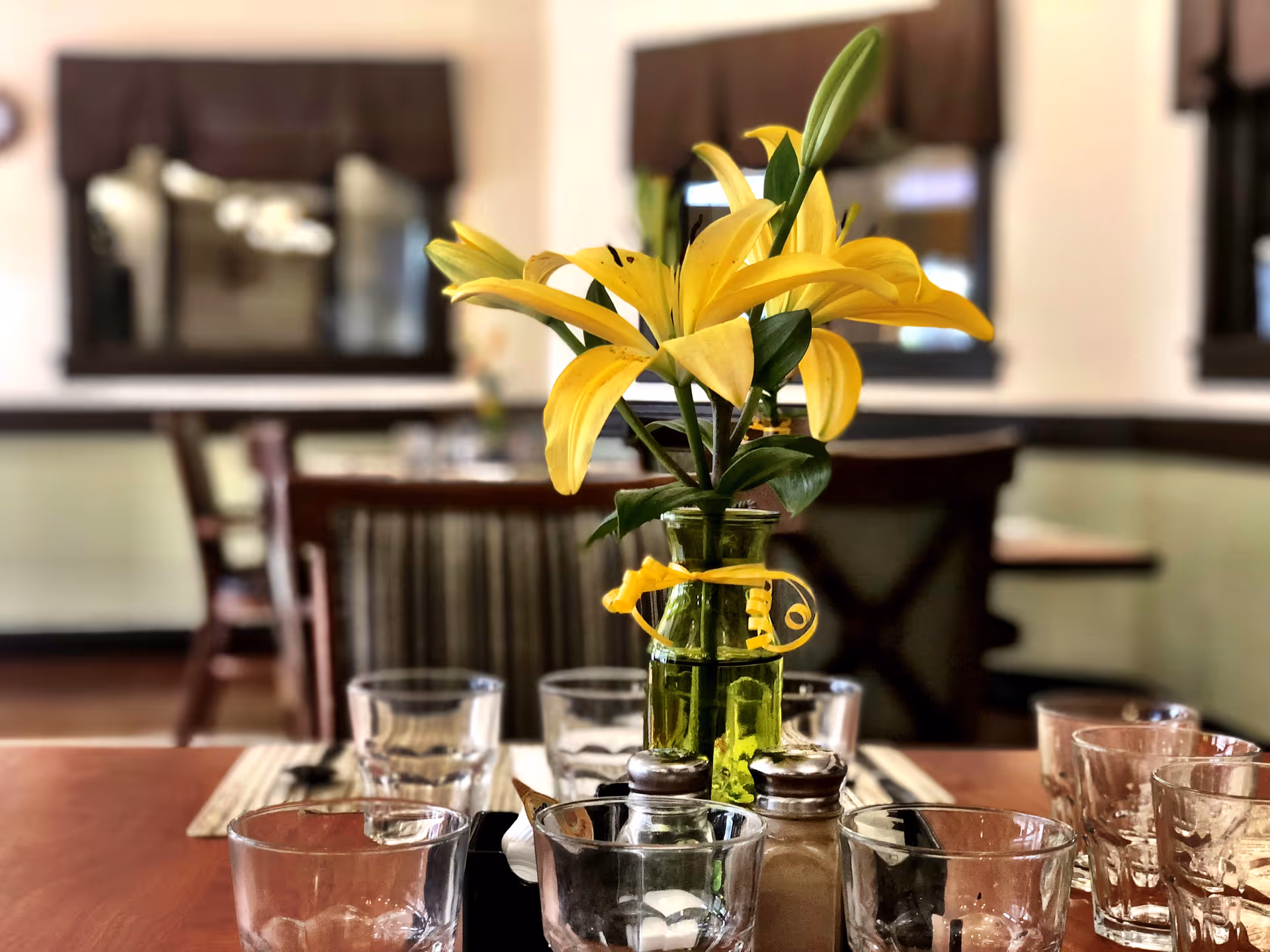 A close-up view of a dining table set with multiple empty drinking glasses, salt and pepper shakers, and a green vase holding yellow lilies. The background shows blurred dining chairs and windows with dark curtains in a softly lit room.