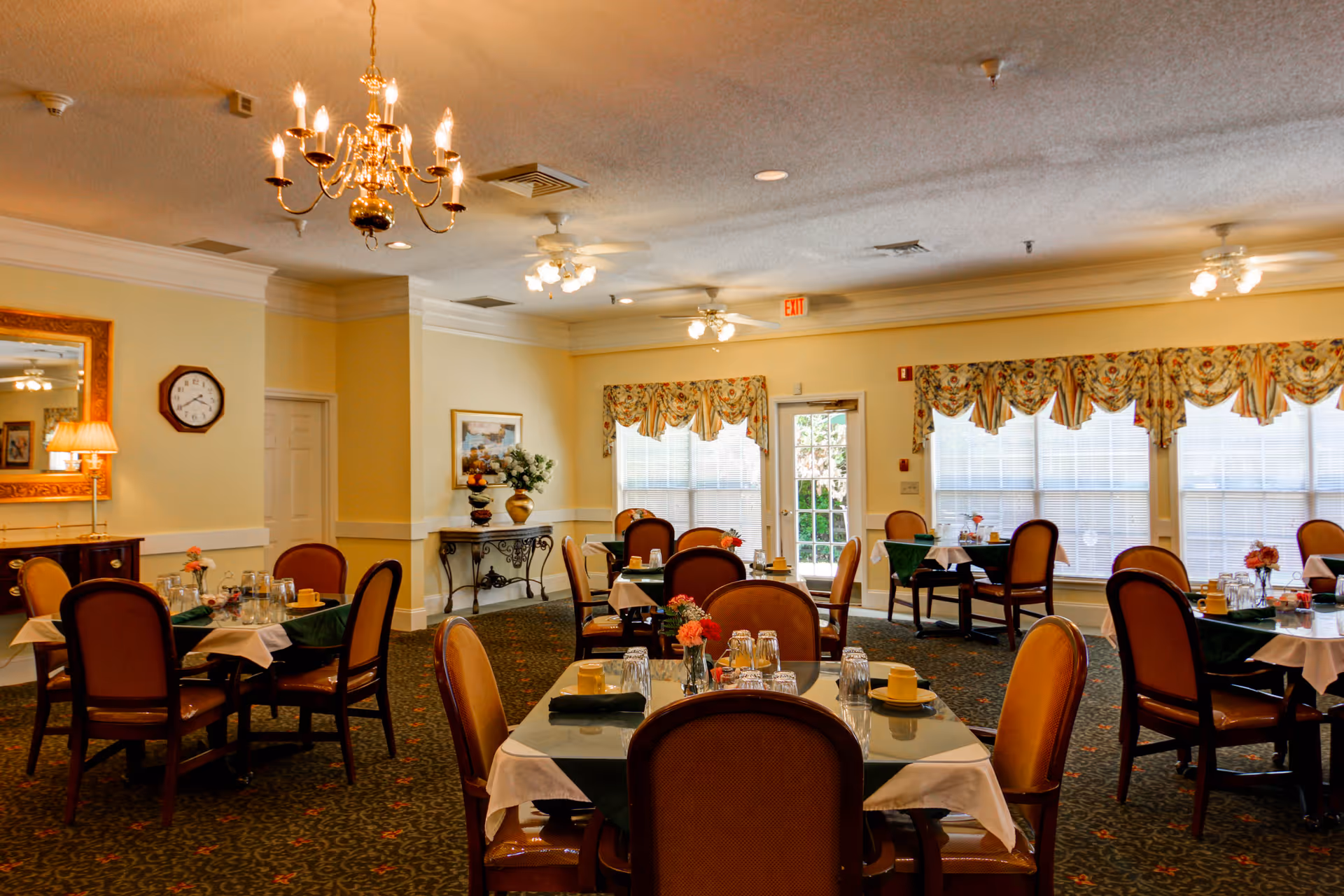 A warmly lit dining room with multiple tables set with tablecloths, napkins, glasses, and small flower arrangements. The room features upholstered chairs, a chandelier, ceiling fans, large windows with floral valances, a wall clock, a mirror, and decorative plants on a console table.