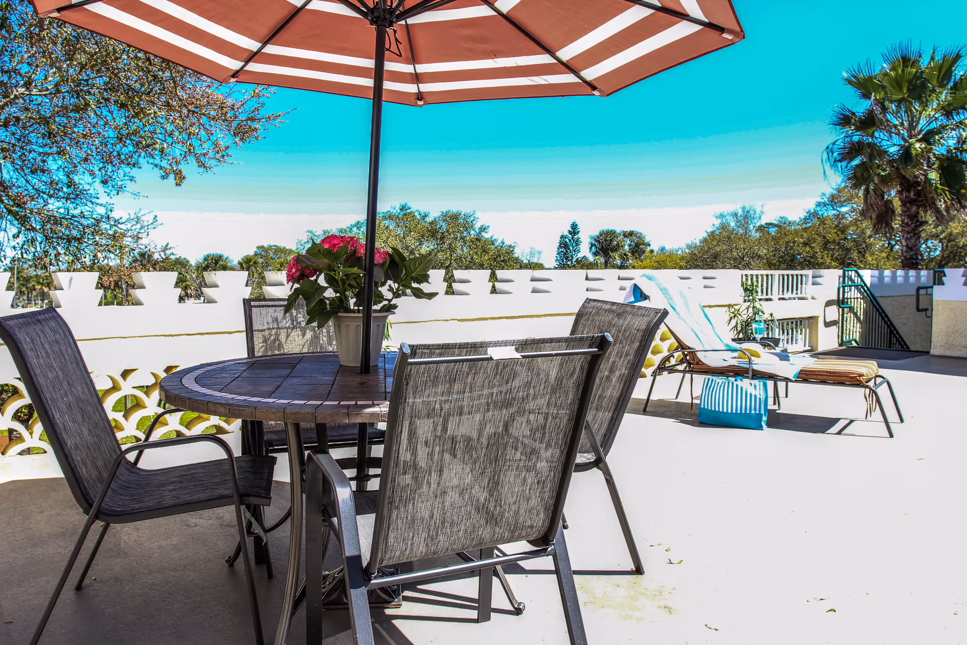 Sunlit outdoor patio with a table and umbrella, several chairs, lounge chairs, and a potted plant overlooking trees.