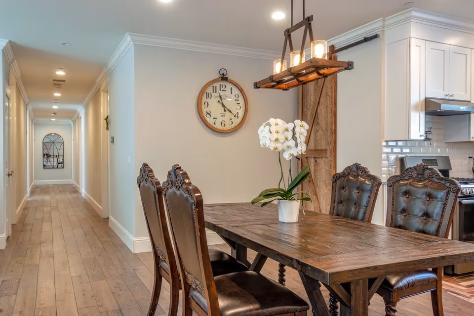 A dining area with a wooden table and four ornate dark leather chairs. A white orchid plant in a pot is placed on the table. Above the table hangs a rectangular light fixture with exposed bulbs. On the wall behind the table is a round clock with a wooden frame. To the right, part of a kitchen with white cabinets and a stainless steel stove is visible. A long hallway with wooden flooring and white walls extends to the left, ending with a decorative mirror on the far wall.