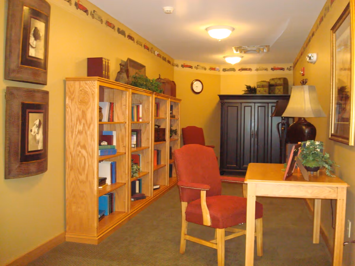 Warmly lit sitting area with bookshelves, a wooden desk and red upholstered chairs in a small common room.