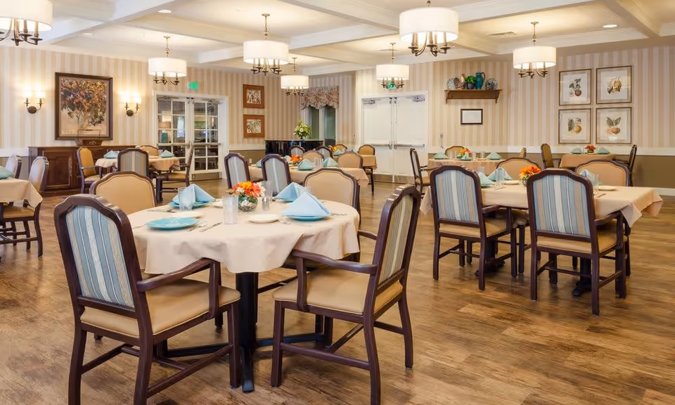 A well-lit dining room with multiple round tables covered with beige tablecloths, each set with blue napkins, plates, and silverware. The room features wooden chairs with cushioned seats and backs, striped wallpaper, framed artwork on the walls, and several ceiling light fixtures. There are double doors and windows in the background, along with decorative shelves and a piano.